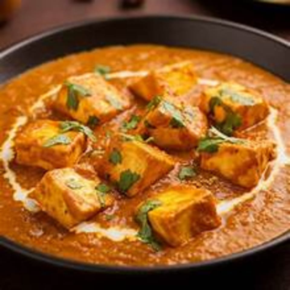 Paneer butter masala, a creamy orange curry with paneer cubes and cilantro in a black bowl.